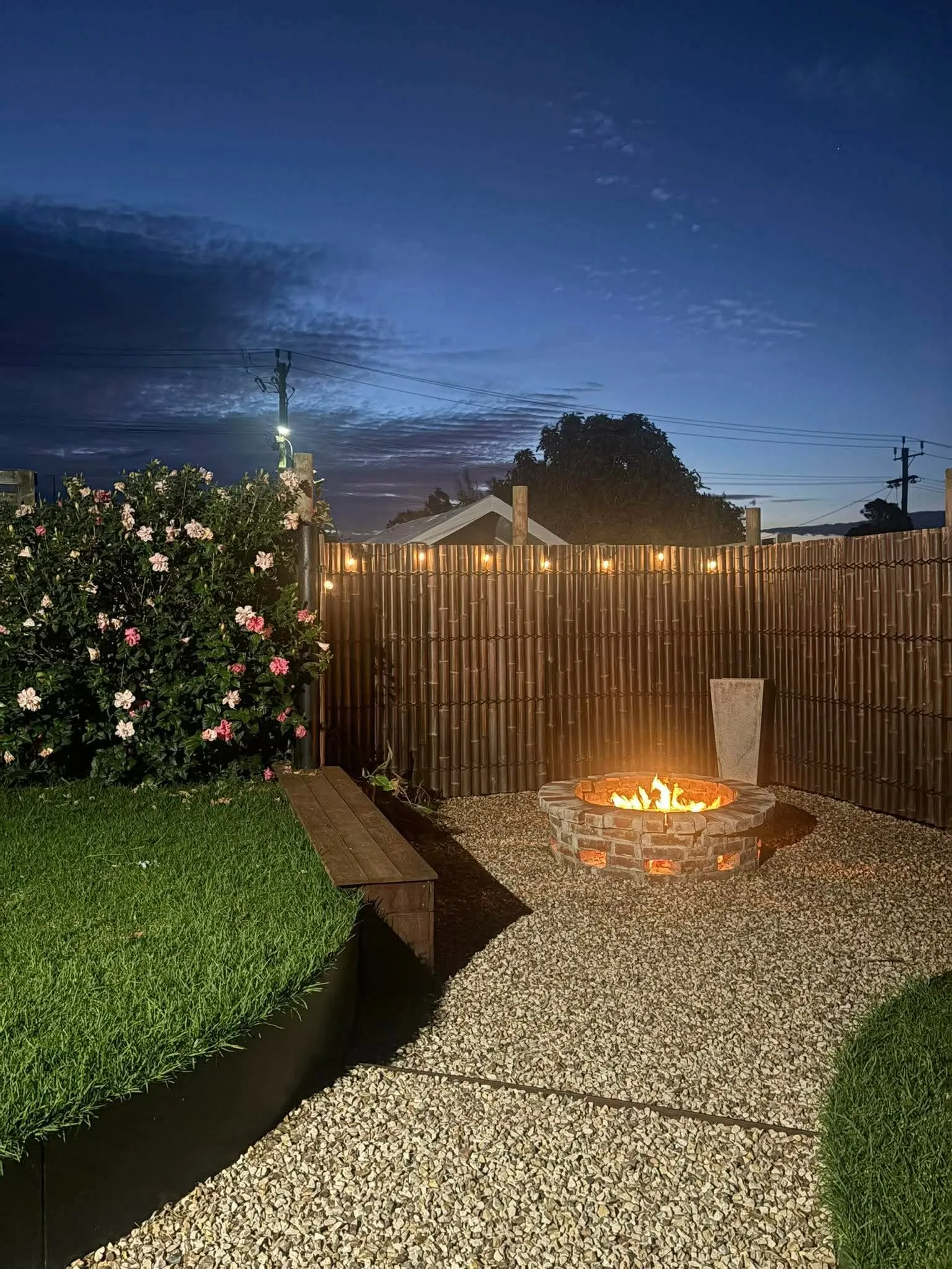 Landscaped backyard at twilight with a brick firepit, gravel and turf areas, steel edging, a bamboo privacy screen, flowering shrub and garden lighting