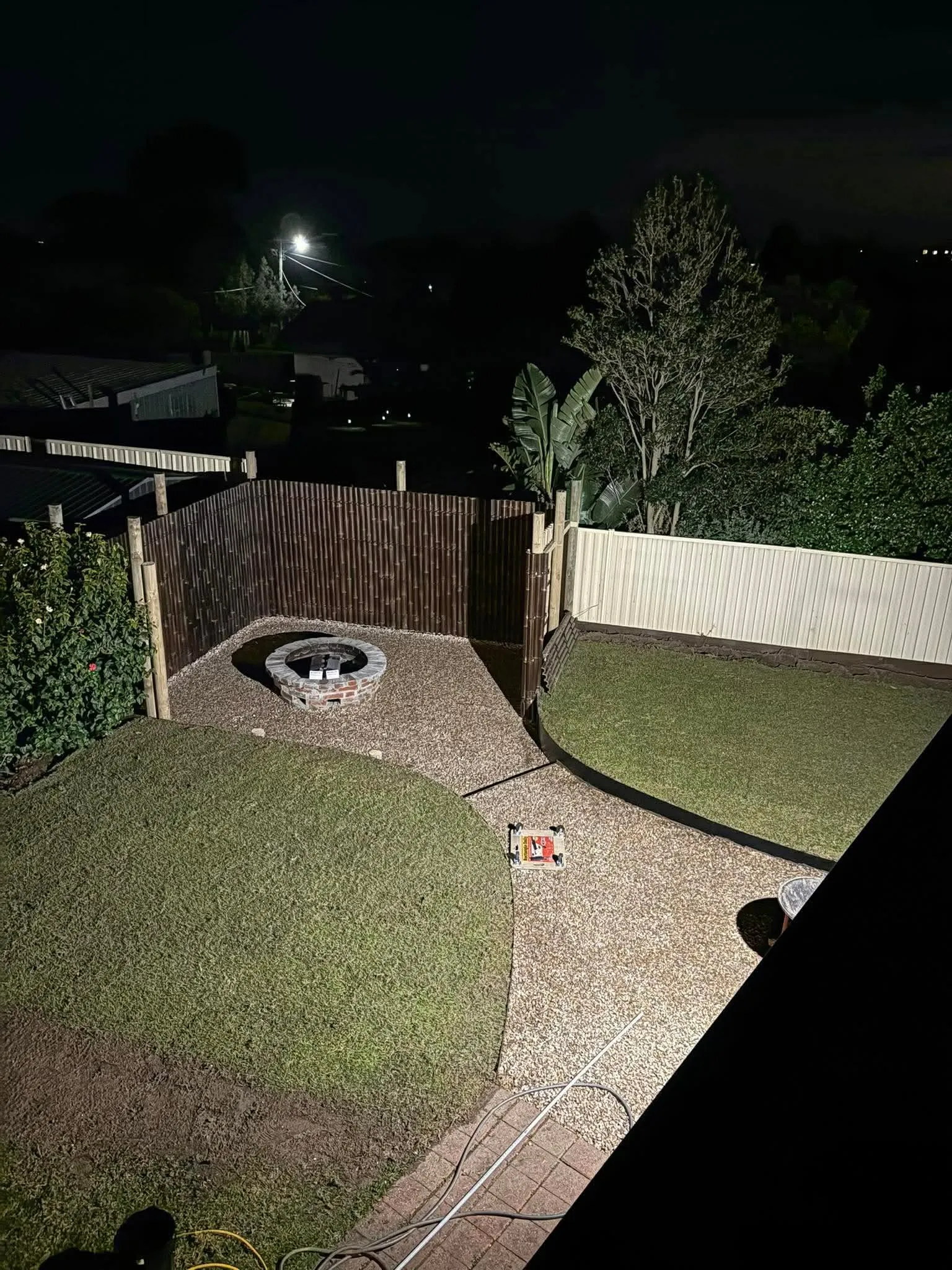 Night view of a landscaped backyard with a brick firepit, shaped turf, gravel walkway and a bamboo privacy screen
