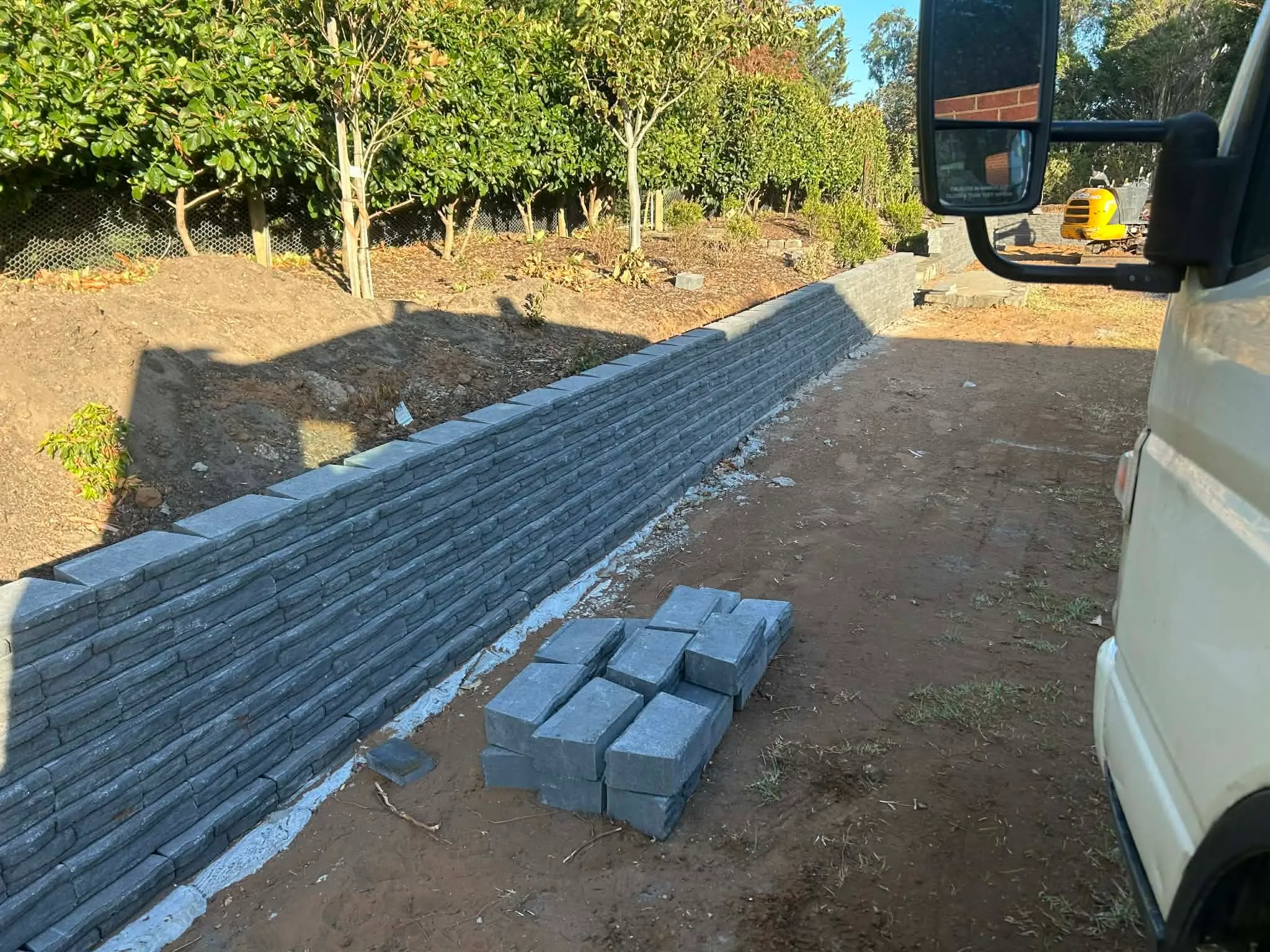 Newly built charcoal concrete-block retaining wall along a sloping boundary, with capping blocks ready to lay