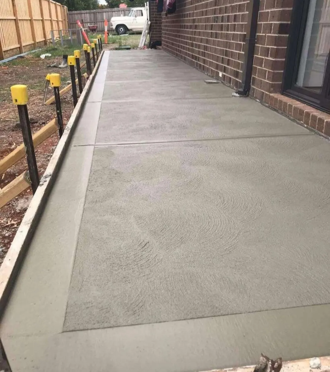 Freshly poured concrete side path with formwork still in place, running between a brick home and timber fence