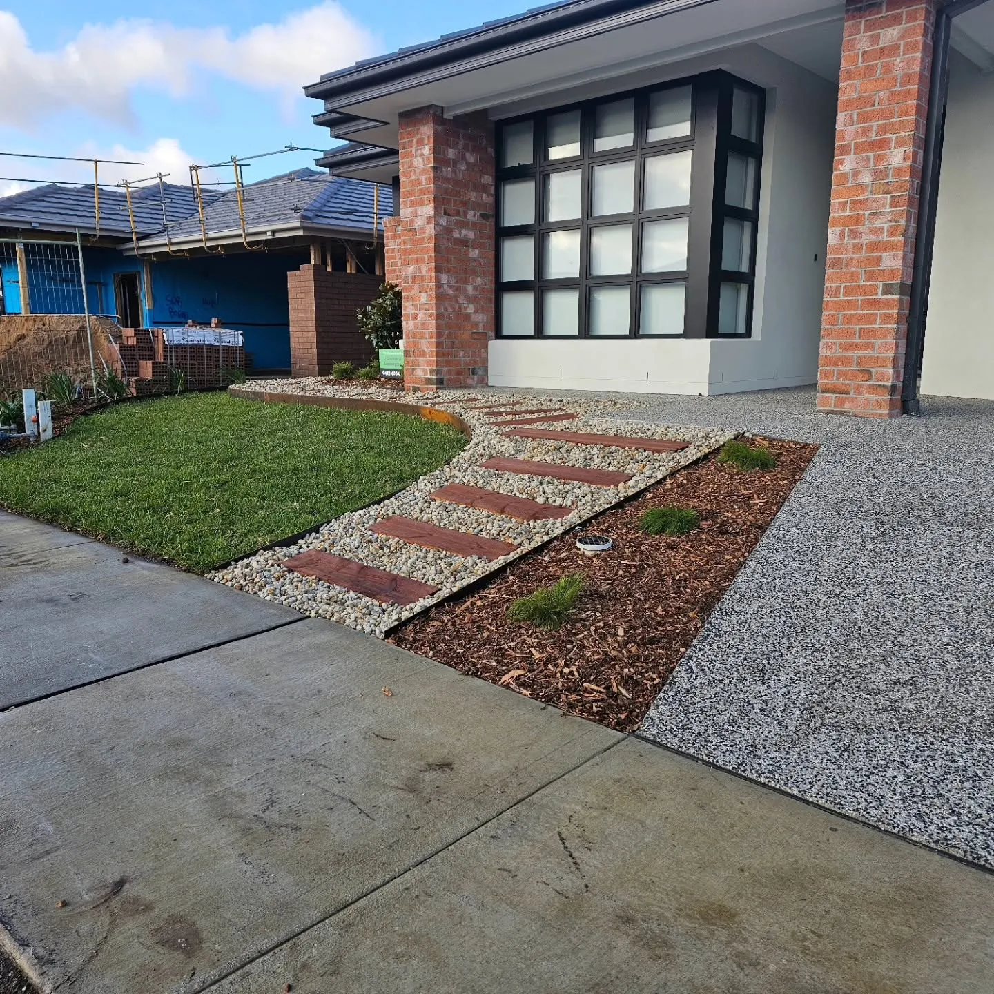 Front yard with a brick-and-paver stepping path, decorative rock, mulched bed and new lawn beside an exposed-aggregate driveway
