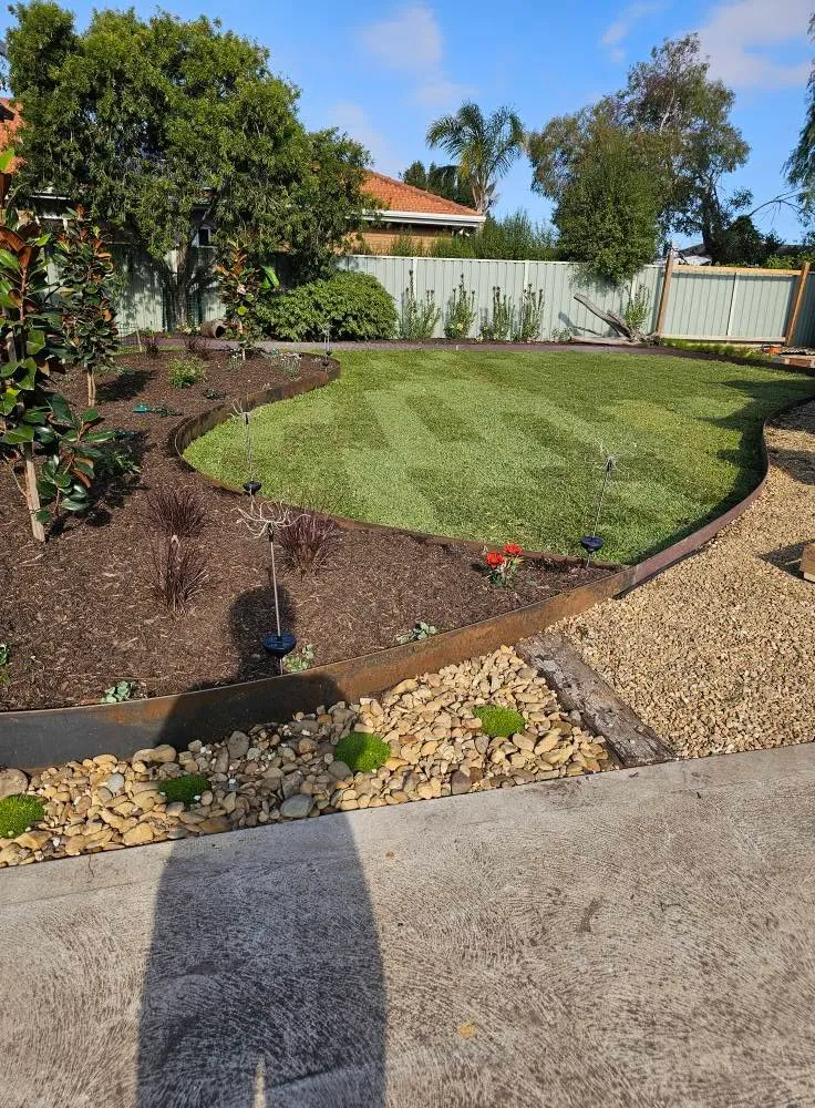 Curved steel-edged garden beds with mulch and shrubs around a freshly mown striped lawn and pebble border